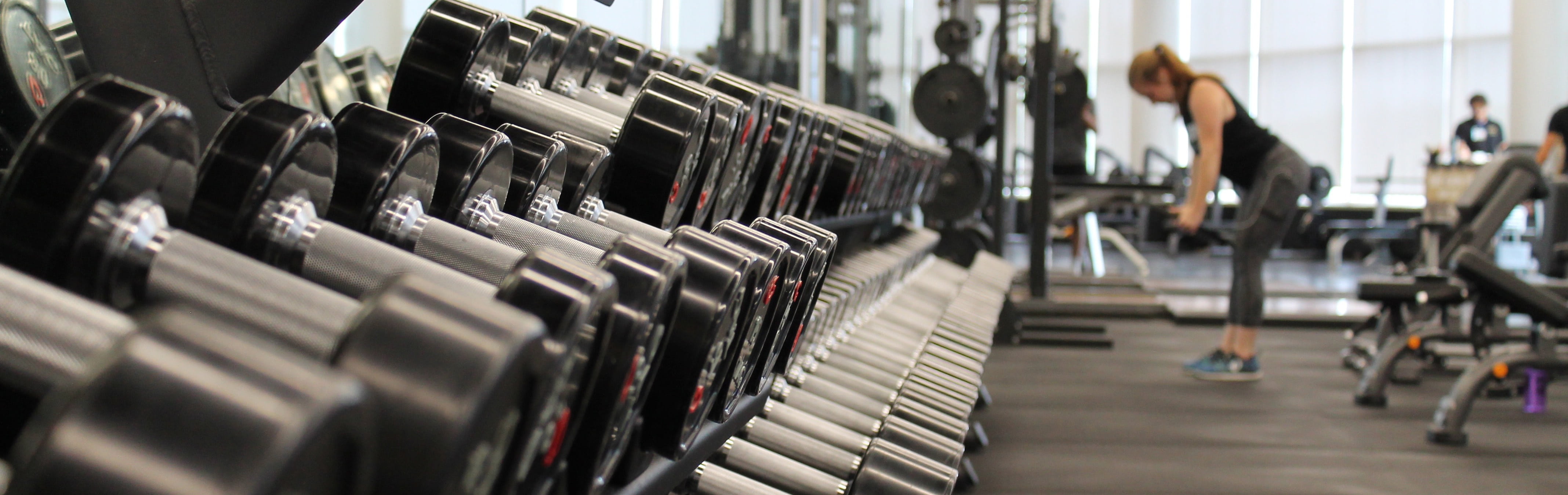 A wide picture of a gym focused on weight rack and in distance a girl doing dumbell rows